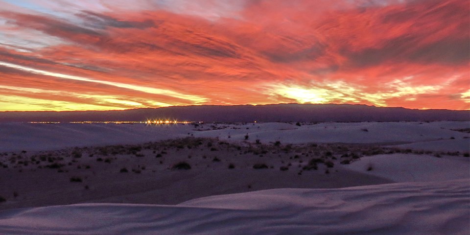 Orange and yellow clouds streak across the sky with white sand dunes dimly lit in the foreground.
