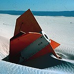 Orange and white rocket mostly embedded in white sand.