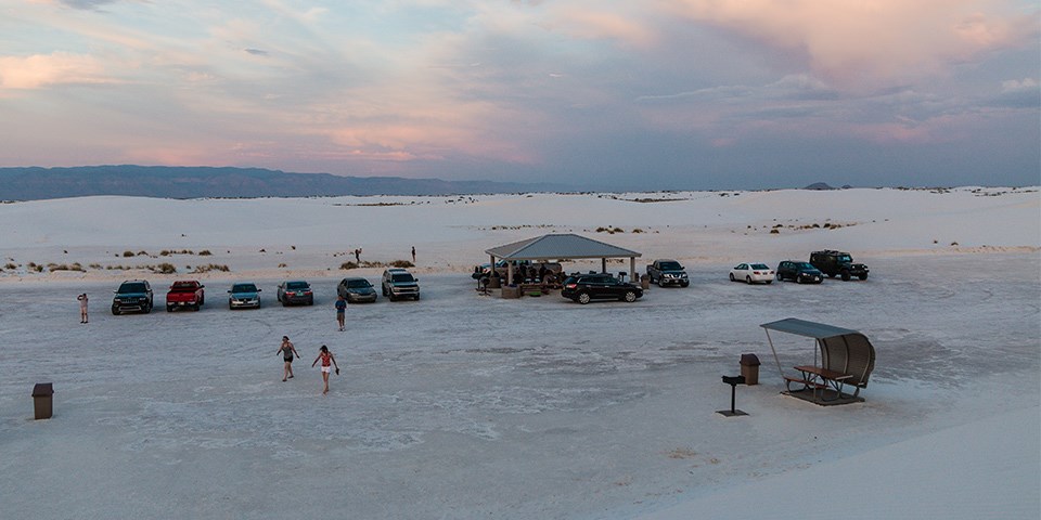 Four small groups of people eating at several picnic tables surrounded by white sand dunes