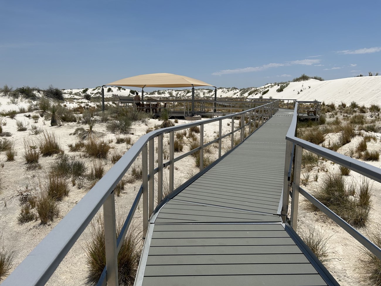 a boardwalk with railings extends into the distance among white sands and sporadic plant life. A covered pavilion is seen in the distance.