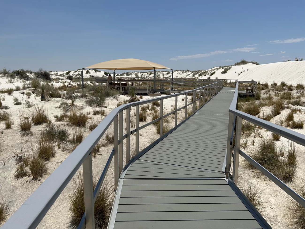 Boardwalk Accessible a boardwalk with railings extends into the distance among white sands and sporadic plant life. A covered pavilion is seen in the distance.