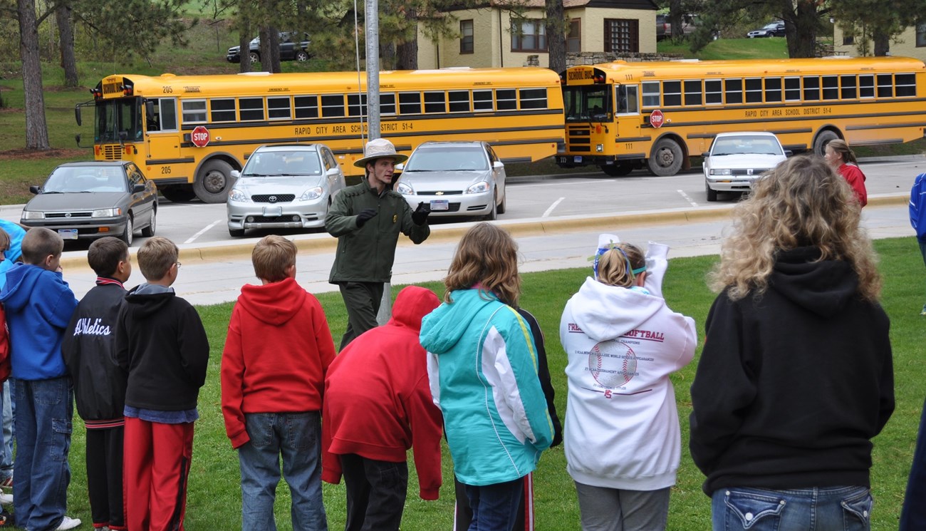 a large group of students standing in a line in front of a park ranger outside, two school buses are parked in the background