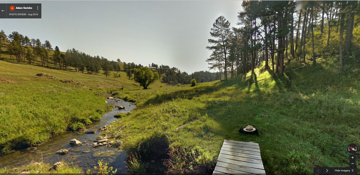 green valley with stream running through it, a ranger flat hat is on the ground adjacent to wooden bridge green valley with stream running through it, a ranger flat hat is on the ground adjacent to wooden bridge