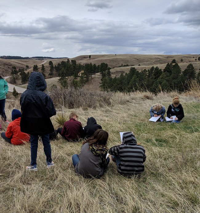Kids sitting on a hillside drawing on clipboards.