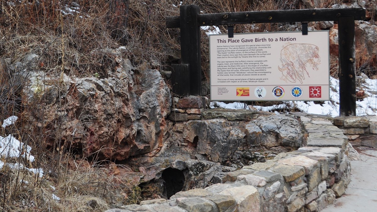 a small hole in a rocky outcrop next to a sign reading "this place gave birth to a nation" with several tribal seals