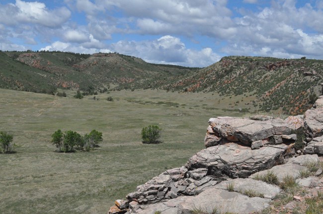 a gray rocky cliff overlooking a wide grassy valley with hills dotted with shrubs on the other side