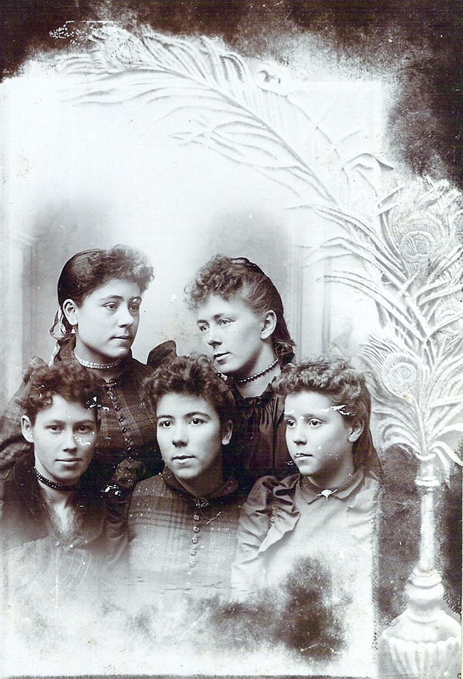 five young women with curly hair and old fashioned dresses pose in a black and white photo