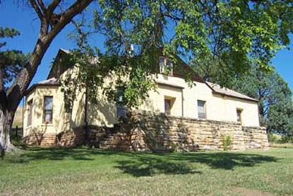 a yellow house with a stone base and brown roof next to a tall leafy tree