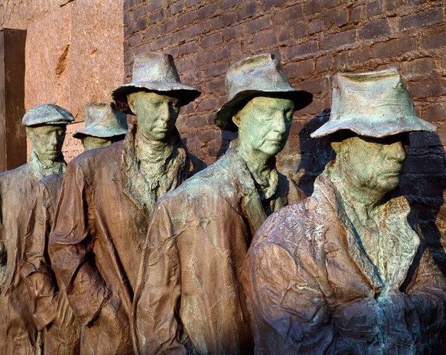 Statues of five men in a line gazing downward. They are wearing hats and tattered coats.