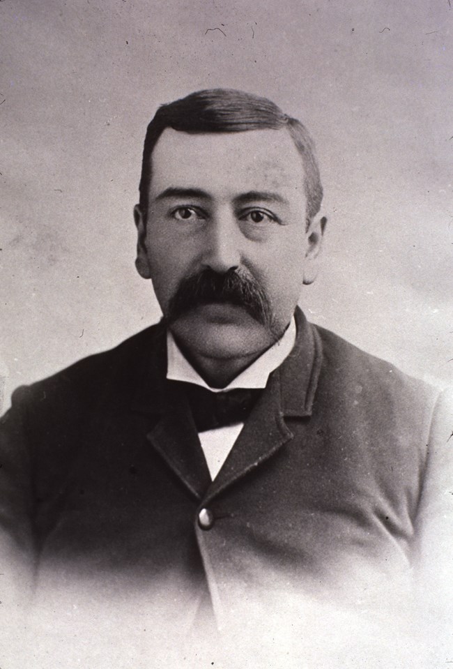 Historic black and white photographic portrait of John Stabler, a white man with a mustache wearing a dark suit and white shirt.