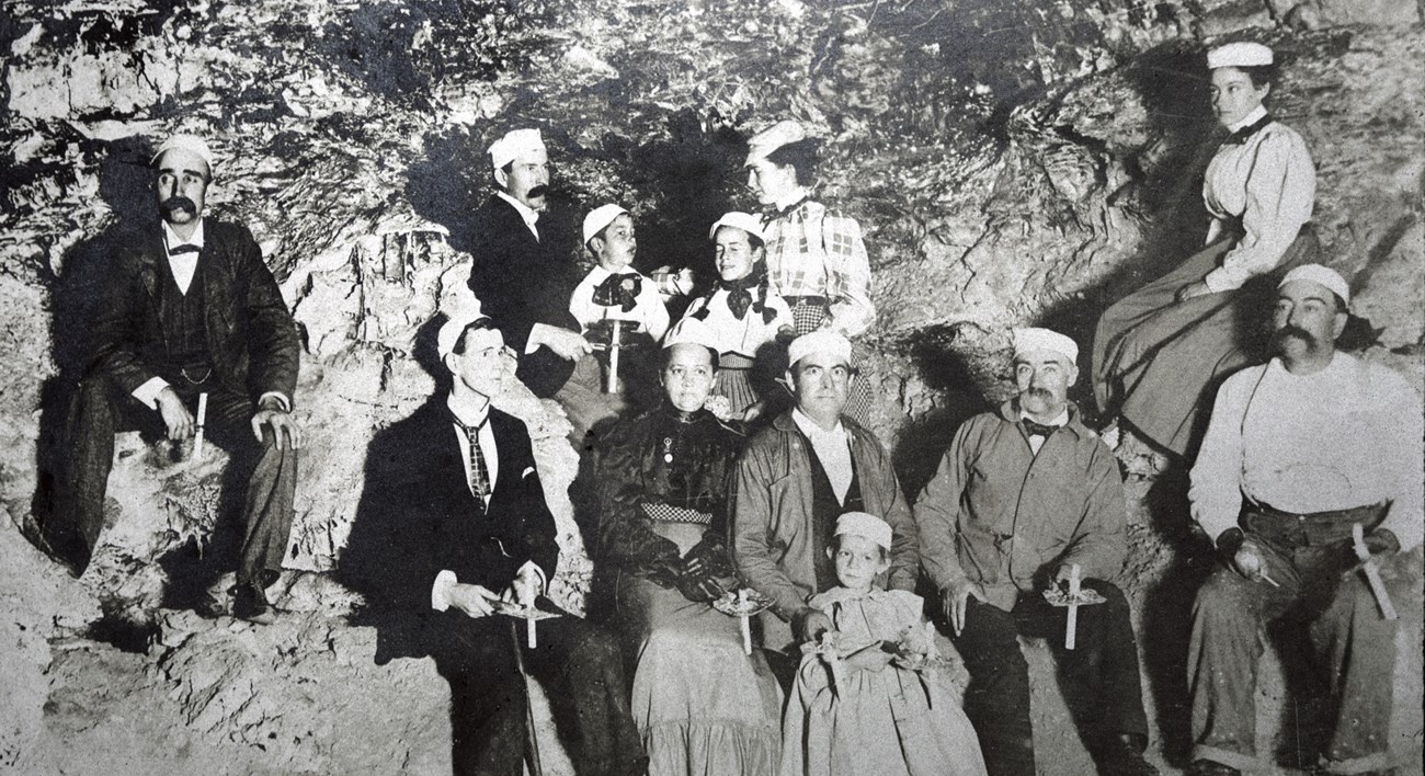black and white photo of a small group of people in clothing from the 1890s holding candles wearing white caps in a cave room