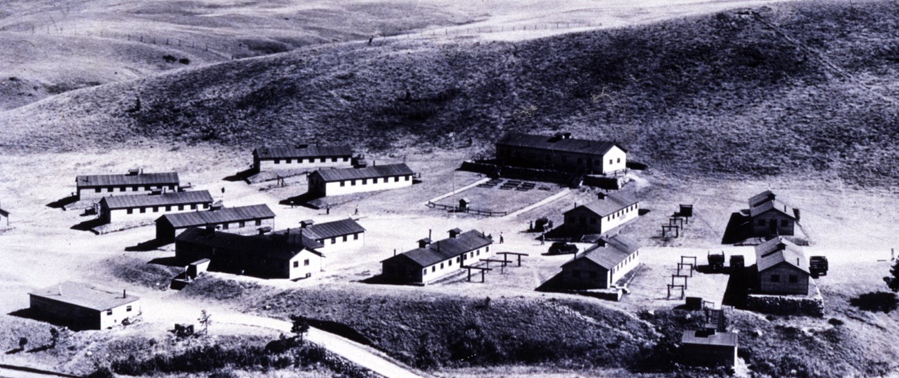 Black and white photo of Camp Wind Cave with 2 rows of 11 barracks arranged in front of a large community building in the background.