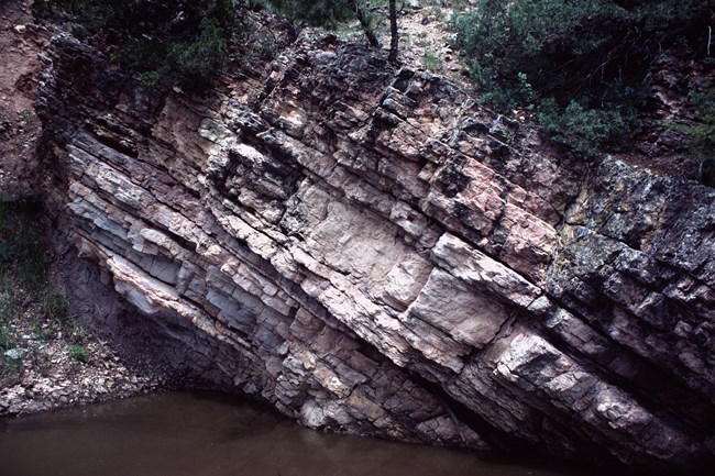 Layers of sedimentary rock titled from upper left to lower right next to a small stream at the bottom.