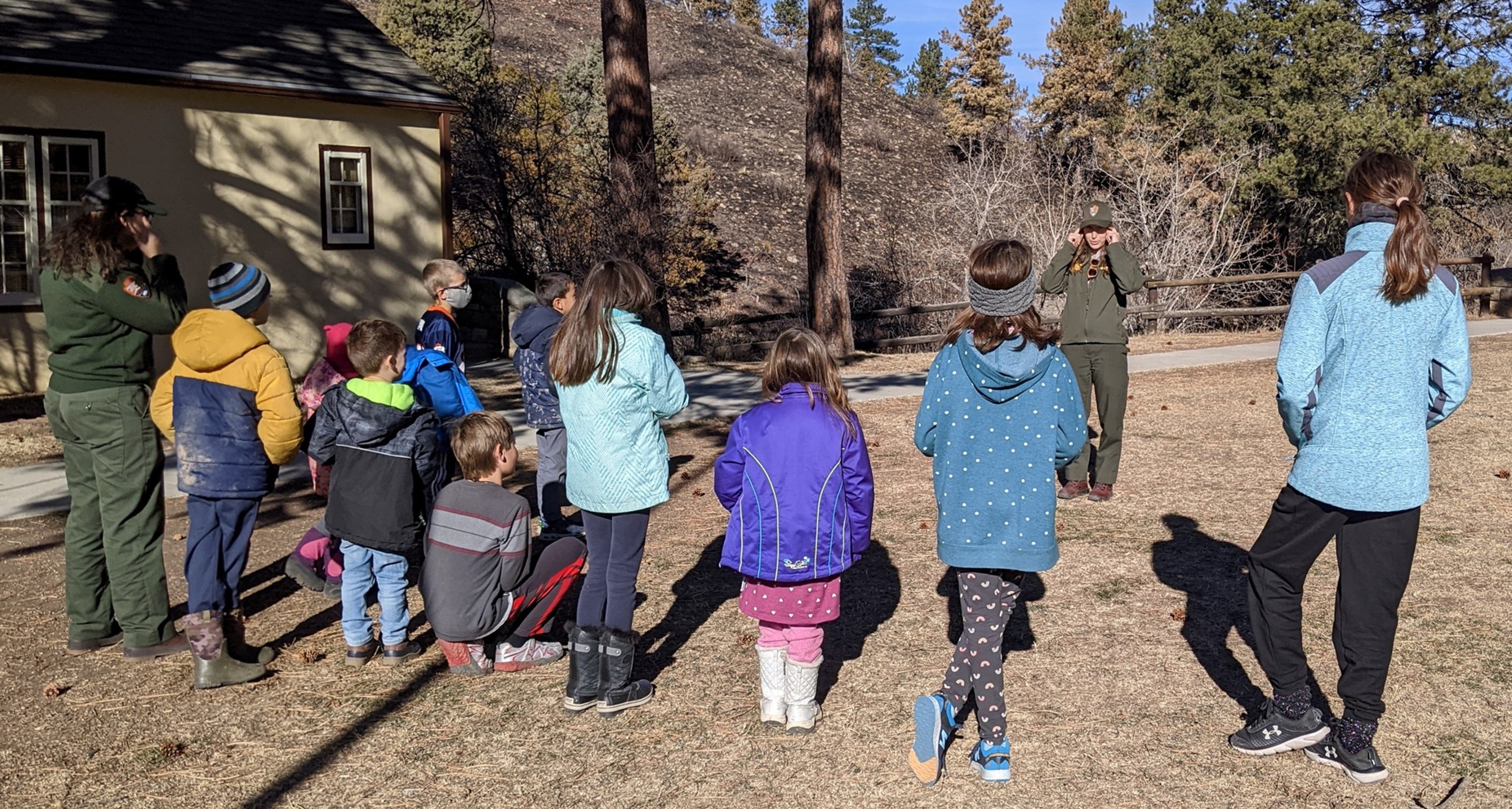 Young children with their backs to the camera dressed in winter coats standing on a brown lawn looking at a ranger in a green uniform who is talking to them. In the background is the corner of a yellow building with trees and a black hillside.