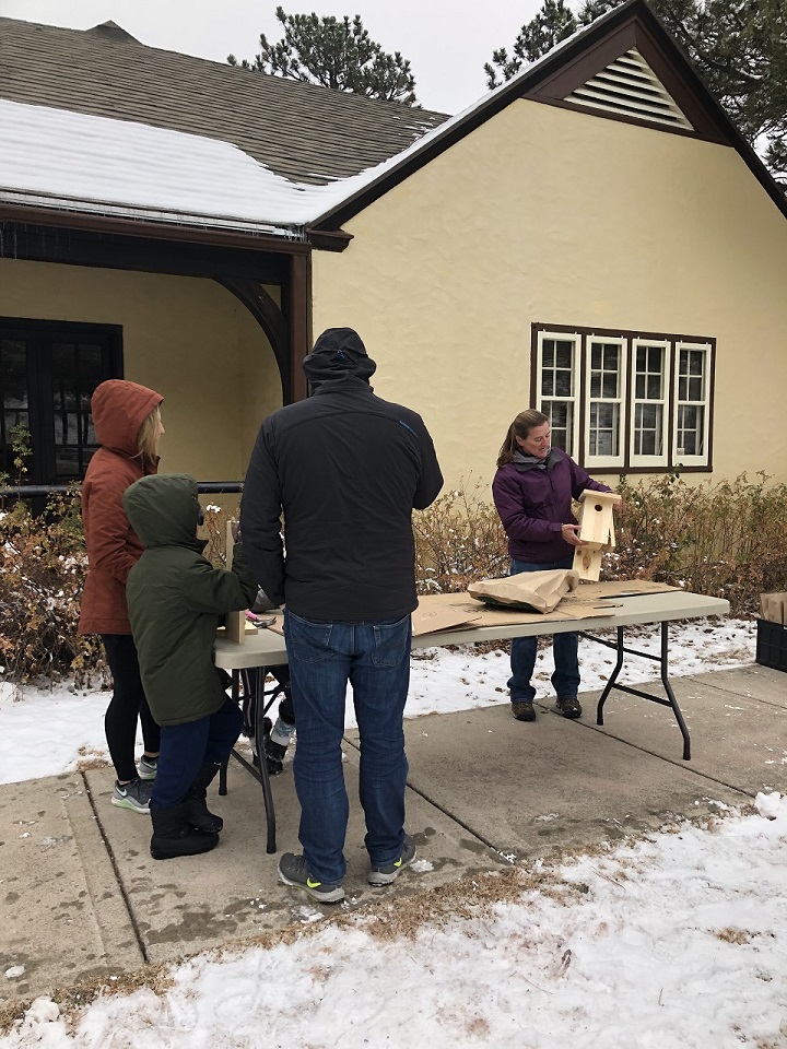 A family of three huddled in winter coats to the photo's left watching a woman in the center of the photo holding a wooden birdbox while standing behind a white table outside with snow on the ground.