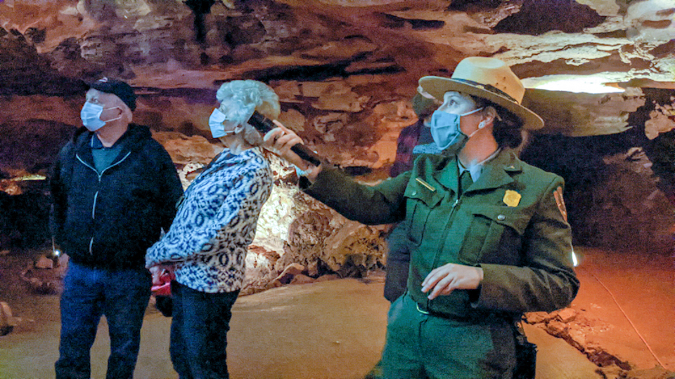 To the right in the photo, a female park ranger is using a flashlight to point to the left while in the background there are two visitors looking in the direction the ranger is pointing. The people are standing in Wind Cave.