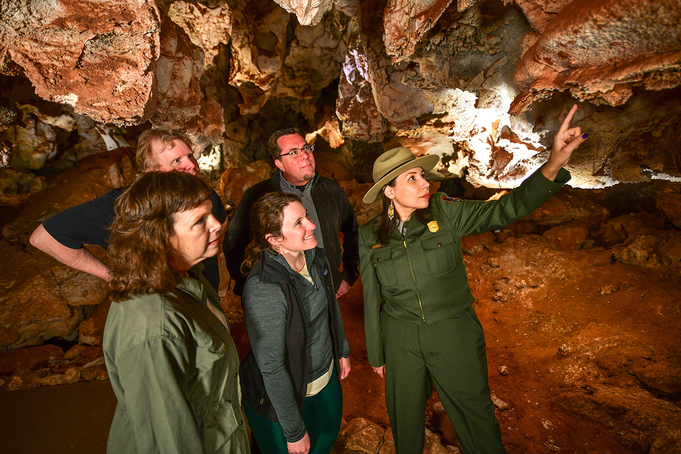 Woman in green park ranger uniform points to a rock in the cave with four visitors behind her looking at where she’s pointing. Behind them the cave is red with areas of white. Woman in green park ranger uniform points to a rock in the cave with four visitors behind her looking at where she’s pointing. Behind them the cave is red with areas of white.