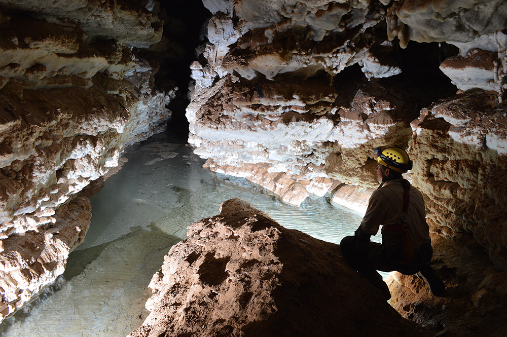 A caver sits by an underground lake. His flash illuminating a body of water in the cave passage in front of him.