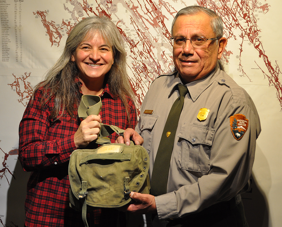 Rachel Cox hands Park Superintendent Vidal Dávila the green cave pack she used the day she became lost 30 years ago in Wind Cave. Both are standing in front of a large cave map. Rachel Cox hands Park Superintendent Vidal Dávila the green cave pack she used the day she became lost 30 years ago in Wind Cave. Both are standing in front of a large cave map.