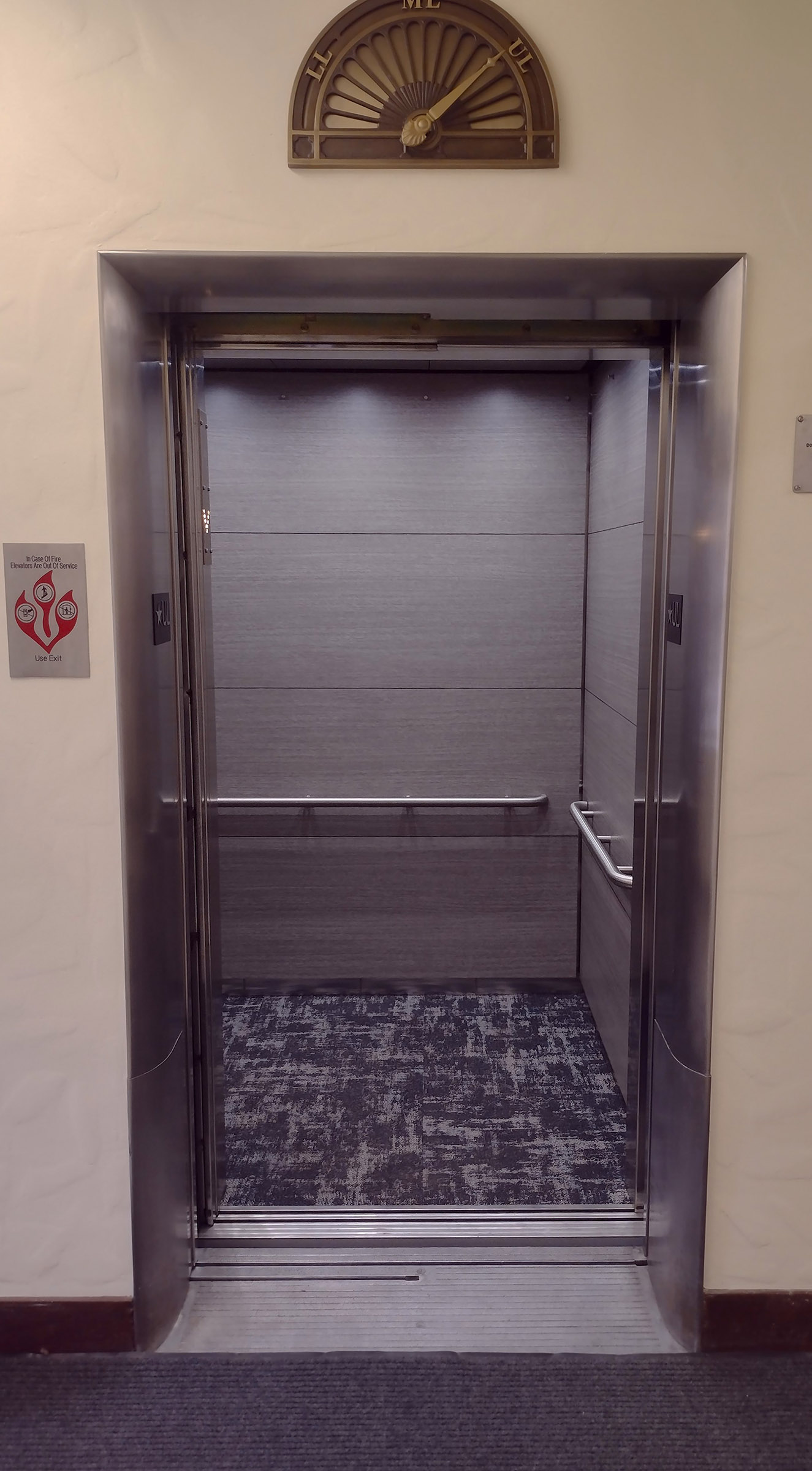 Elevator Entry Looking into an elevator with silver walls and blue and white carpet. There is a bronze dial above the door with an indicator arm.