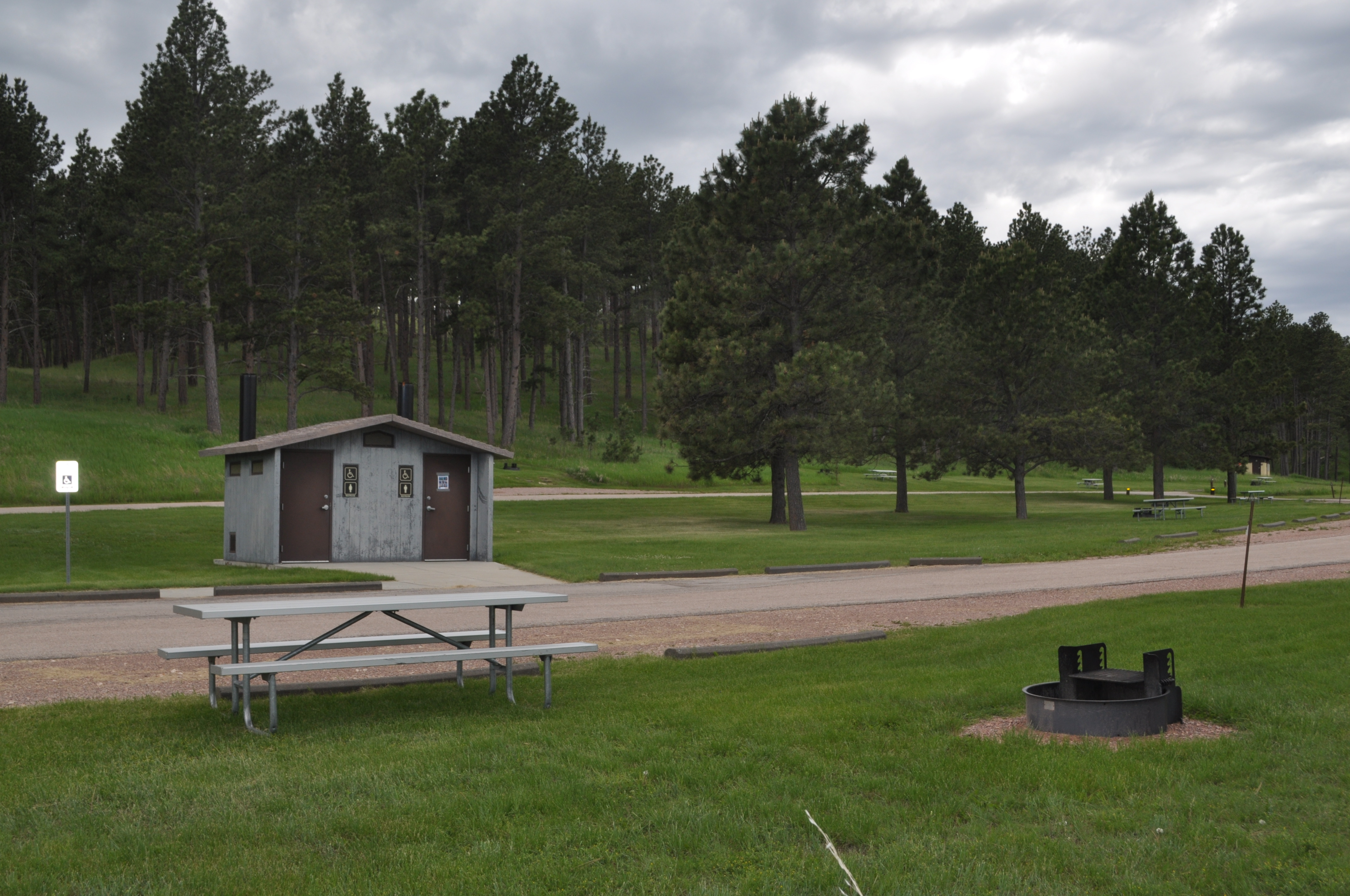 Picnic table on green grass near a fire grate with a vault toilet nearby. There is a slight hill in the background covered with ponderosa pine trees.