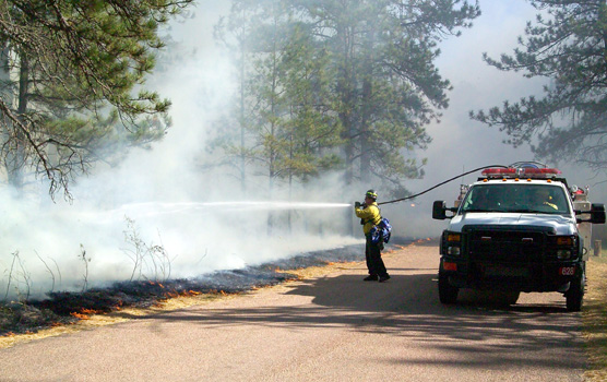 Firefighter spraying water on fire with an engine next to him.