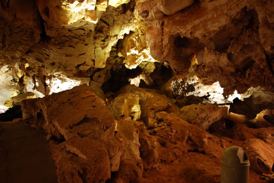 Red rocks piled in a large cave room with a concrete path on the left and small trashcan in front.