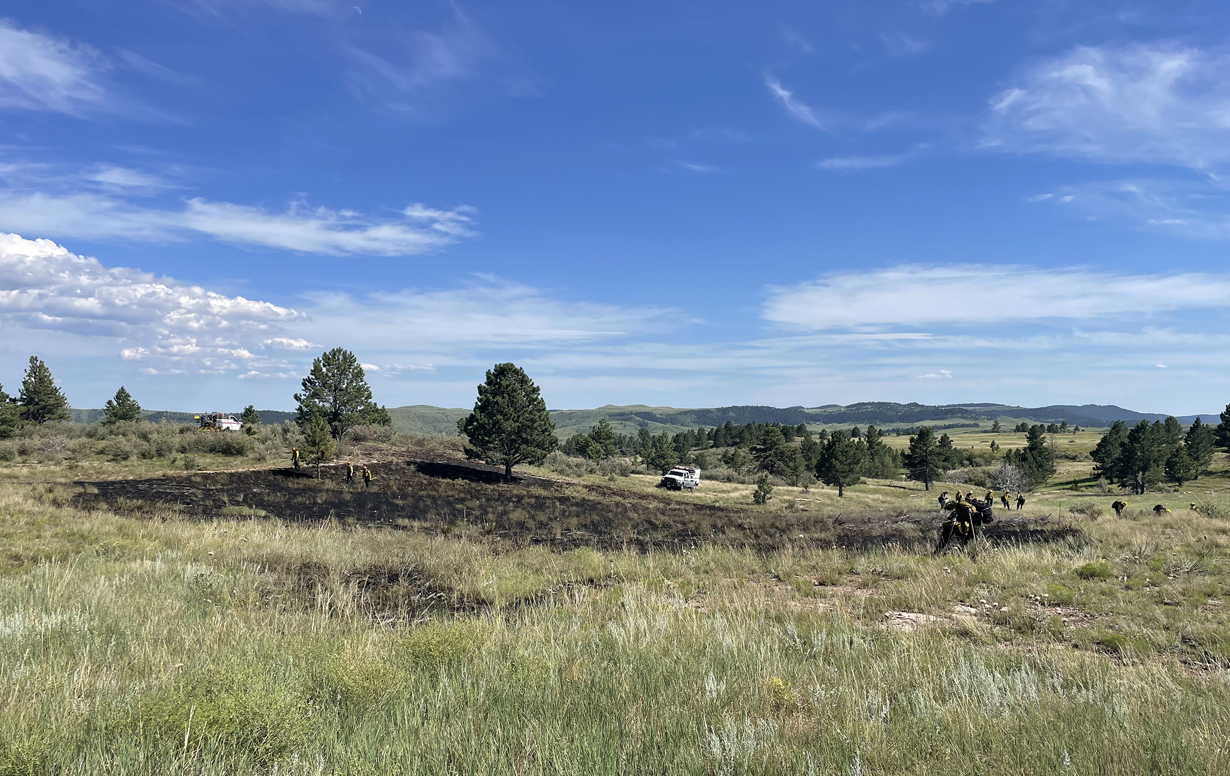 A patch of burnt prairie is in the middle of the photo surrounded by a gently rolling green grass. There are two white fire engines on the far side of the burn with several firefighters working around its perimeter. Blue sky is overhead. A patch of burnt prairie is in the middle of the photo surrounded by a gently rolling green grass. There are two white fire engines on the far side of the burn with several firefighters working around its perimeter. Blue sky is overhead.