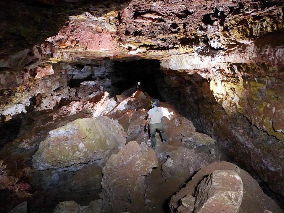 Two cave explorers in a long passageway deep in Wind Cave. Rock walls surround the passage with darkness at the room's far end.