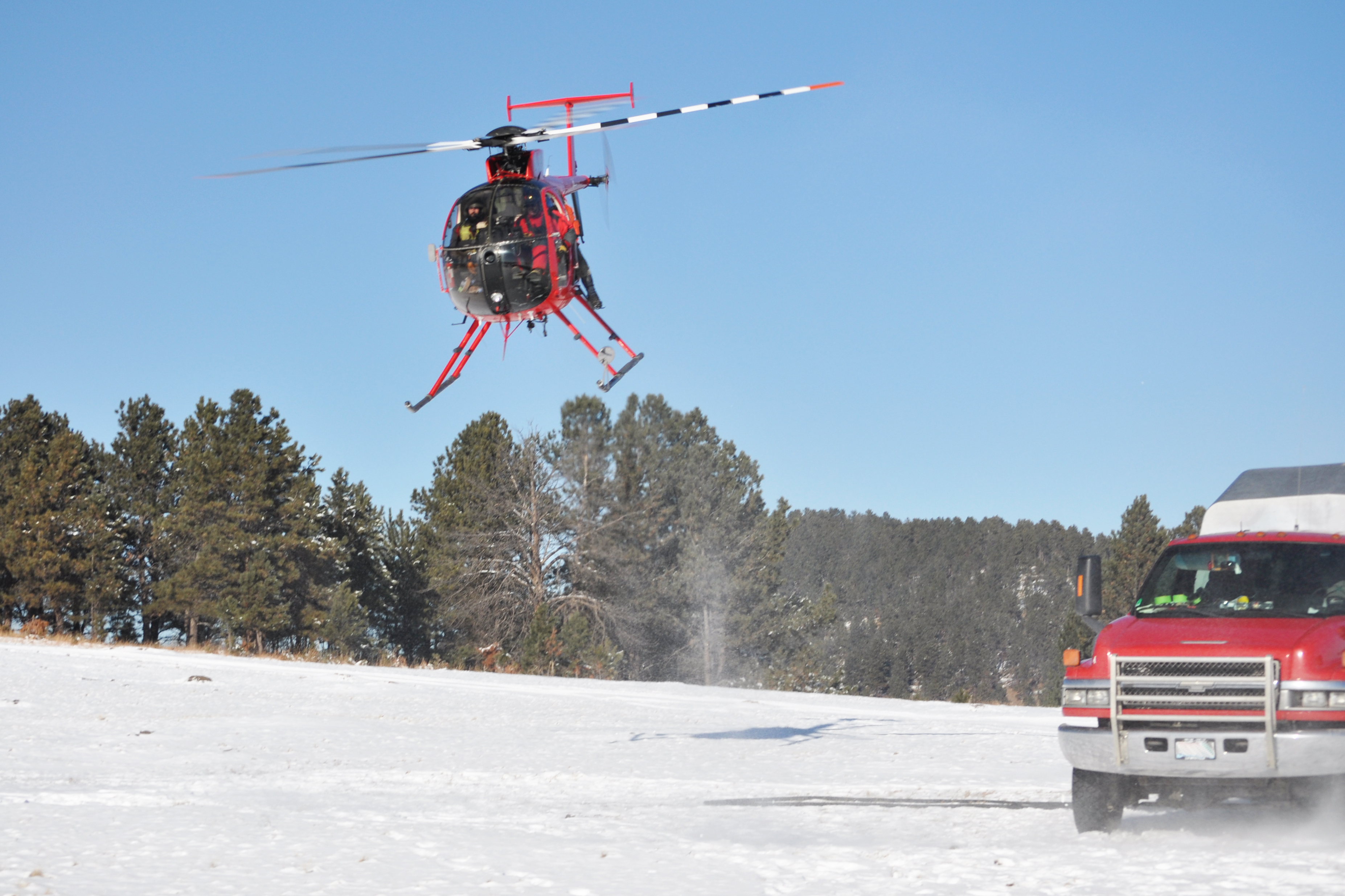 Photo showing a small, red helicopter taking off from a snowy field next to a fuel truck.