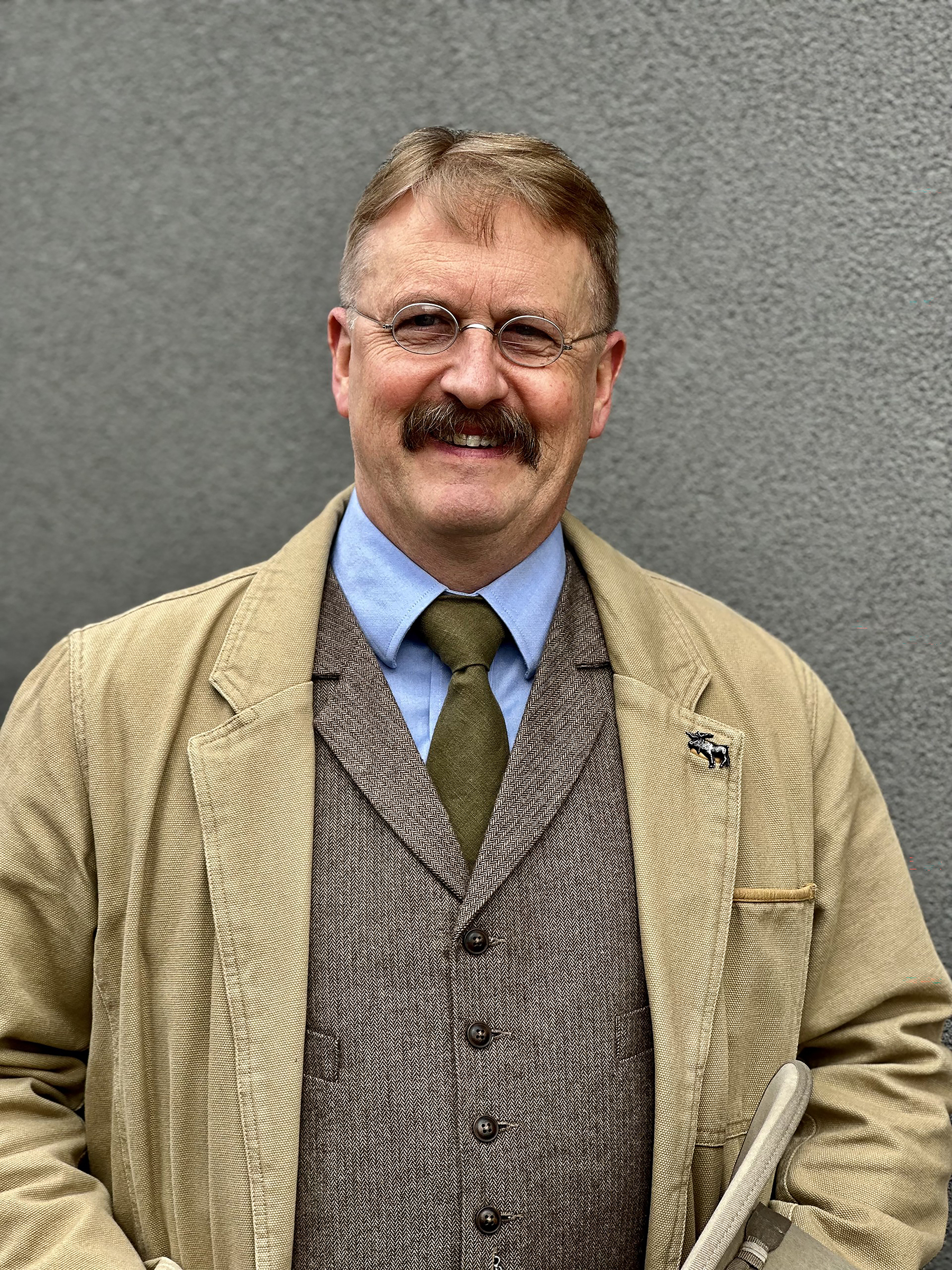 A person with light brown hair, glasses, and a mustache is smiling while standing in front of a gray wall. They are wearing a tan jacket over a brown vest, a blue shirt, and an olive green tie. A small animal-shaped pin is attached to the jacket lapel.