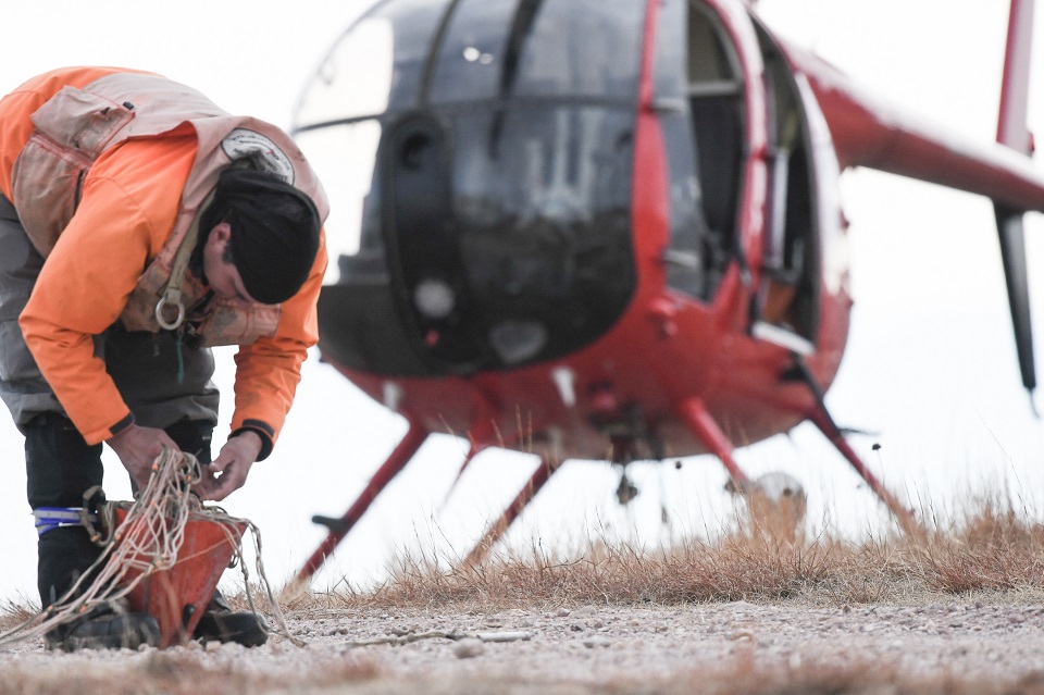 Man in flight gear leaning over and repacking a net into an orange container with a helicopter sitting on the ground behind him.