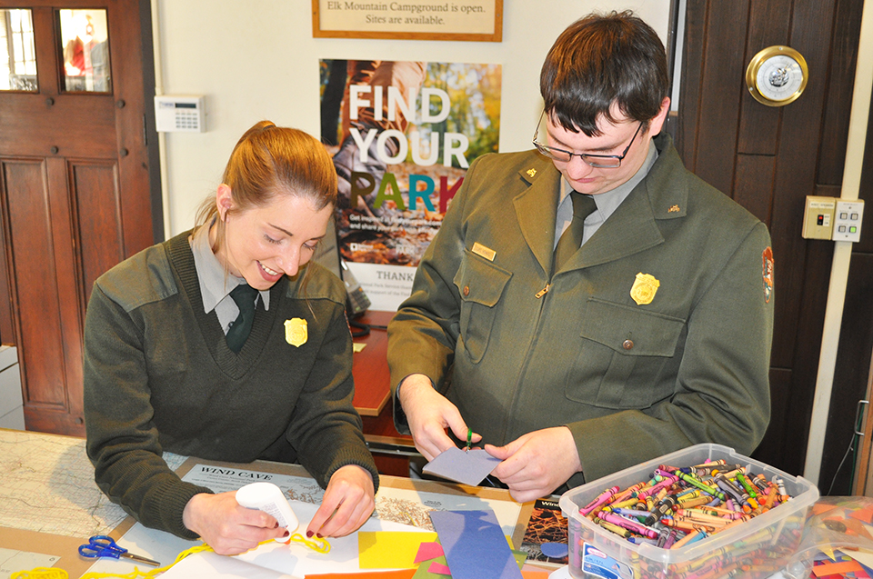 Two rangers stand behind an information desk working on cutting up and gluing colored paper.