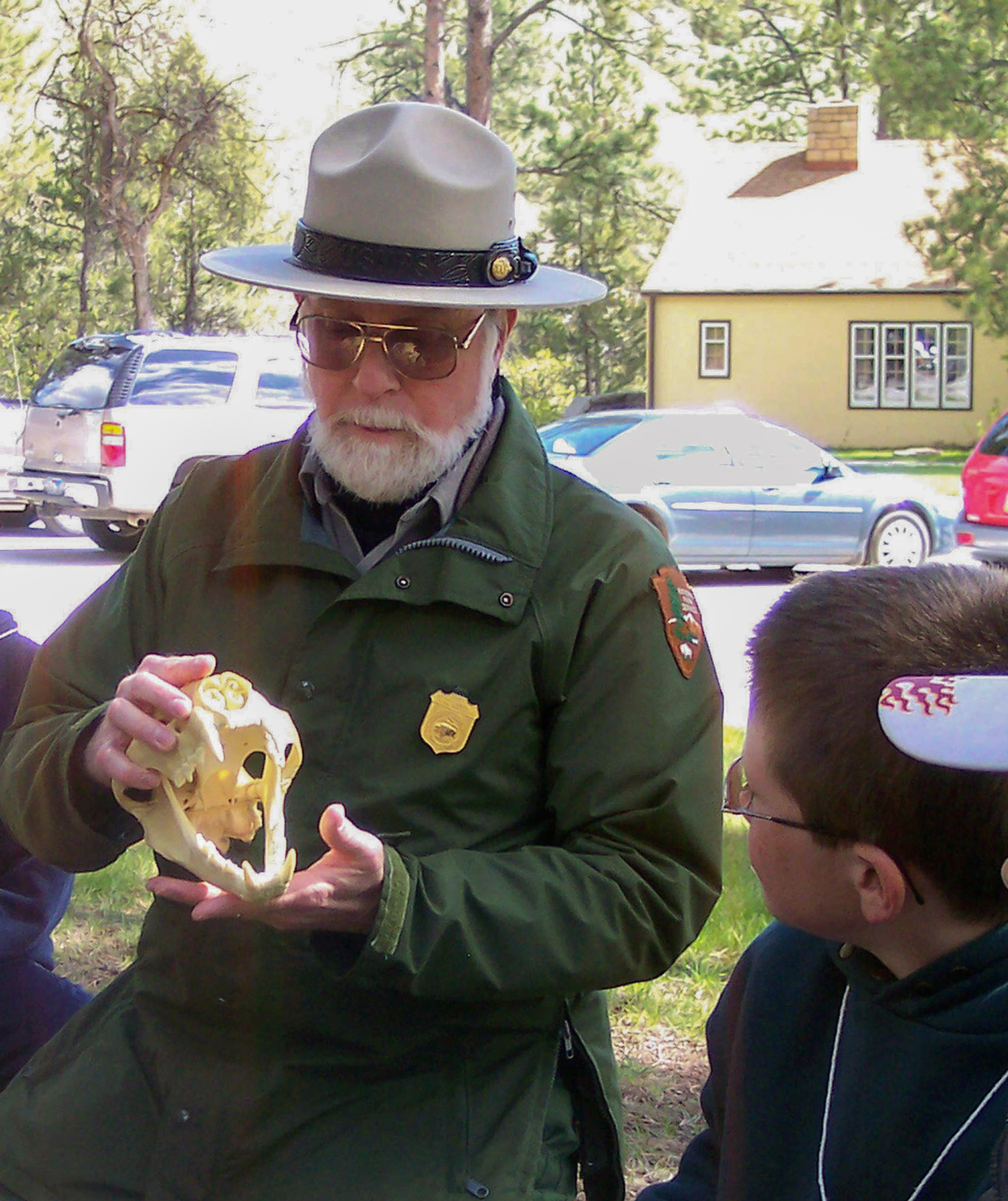Man with a white beard wearing a green coat and ranger hat sits on a lawn holding a plastic model of an animal skull. A young student next to him dressed in dark clothing looks at the skull. Man with a white beard wearing a green coat and ranger hat sits on a lawn holding a plastic model of an animal skull. A young student next to him dressed in dark clothing looks at the skull.