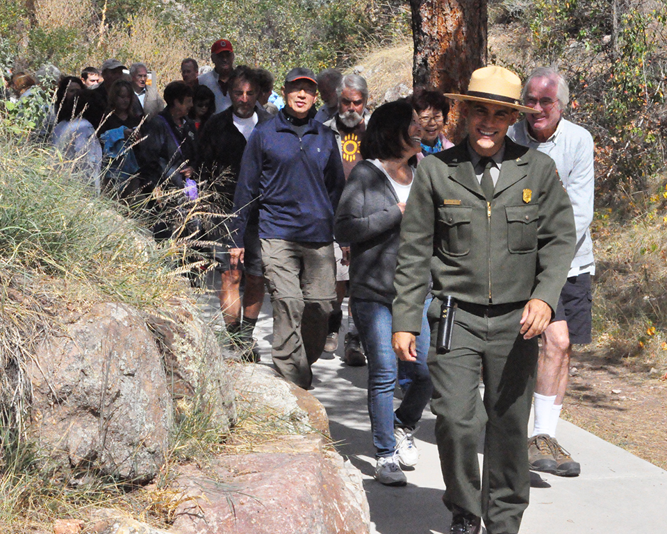 It’s a sunny summer day with a ranger walking toward the camera and 40 people following him. The ranger is walking on a concrete sidewalk and passing by a rock wall to the left of the photo.