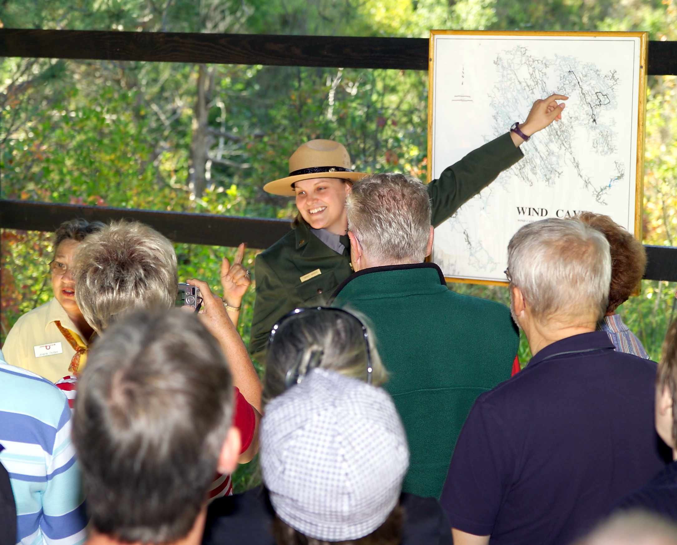 Woman ranger pointing to a map in front of a crowd of people who are watching her with their backs to the camera. She is wearing a green uniform and a ranger hat.