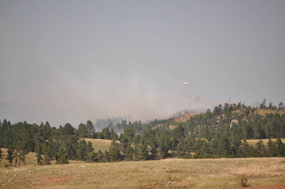 Rankin Fire 2020 A small white plane flies over smoke rising from a tree covered ridge several miles away.
