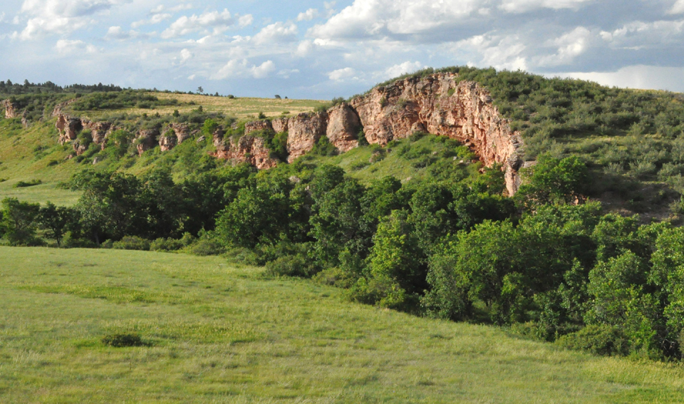 Sanson Buffalo Jump seen from a distance. Rock cliff towering over a green valley.