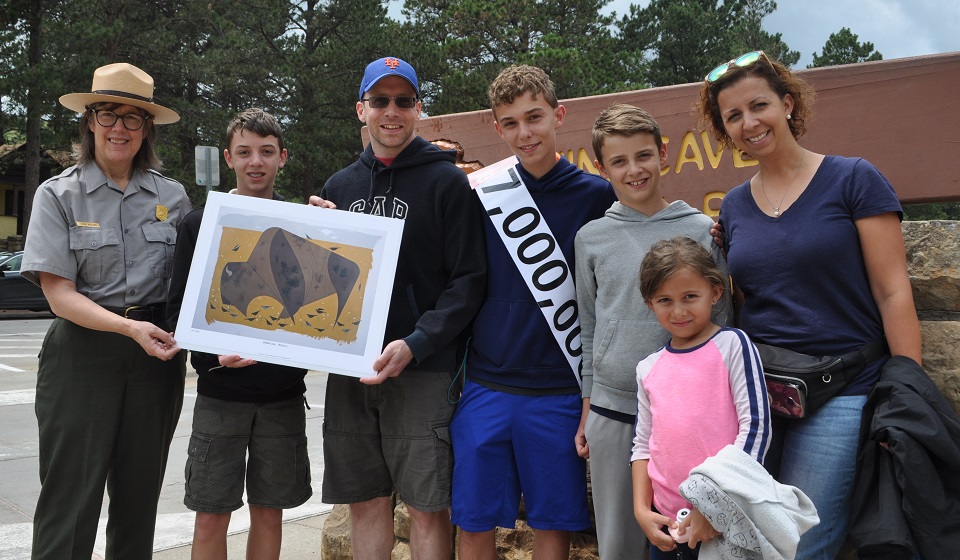 A park ranger, to the left, giving a print of a bison to a family of two adults and four children all standing outside in front of a wooden sign. One of the children wears a sash with “7,000,000” on it.