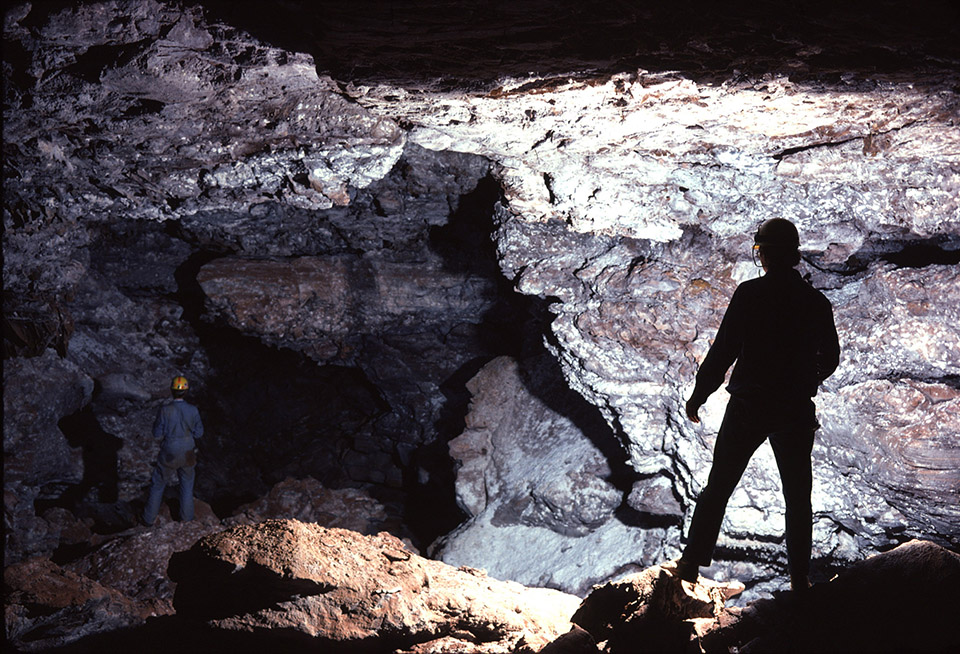Two cave explores stand in a cave room that leads off into the darkness. Two cave explores stand in a cave room that leads off into the darkness.