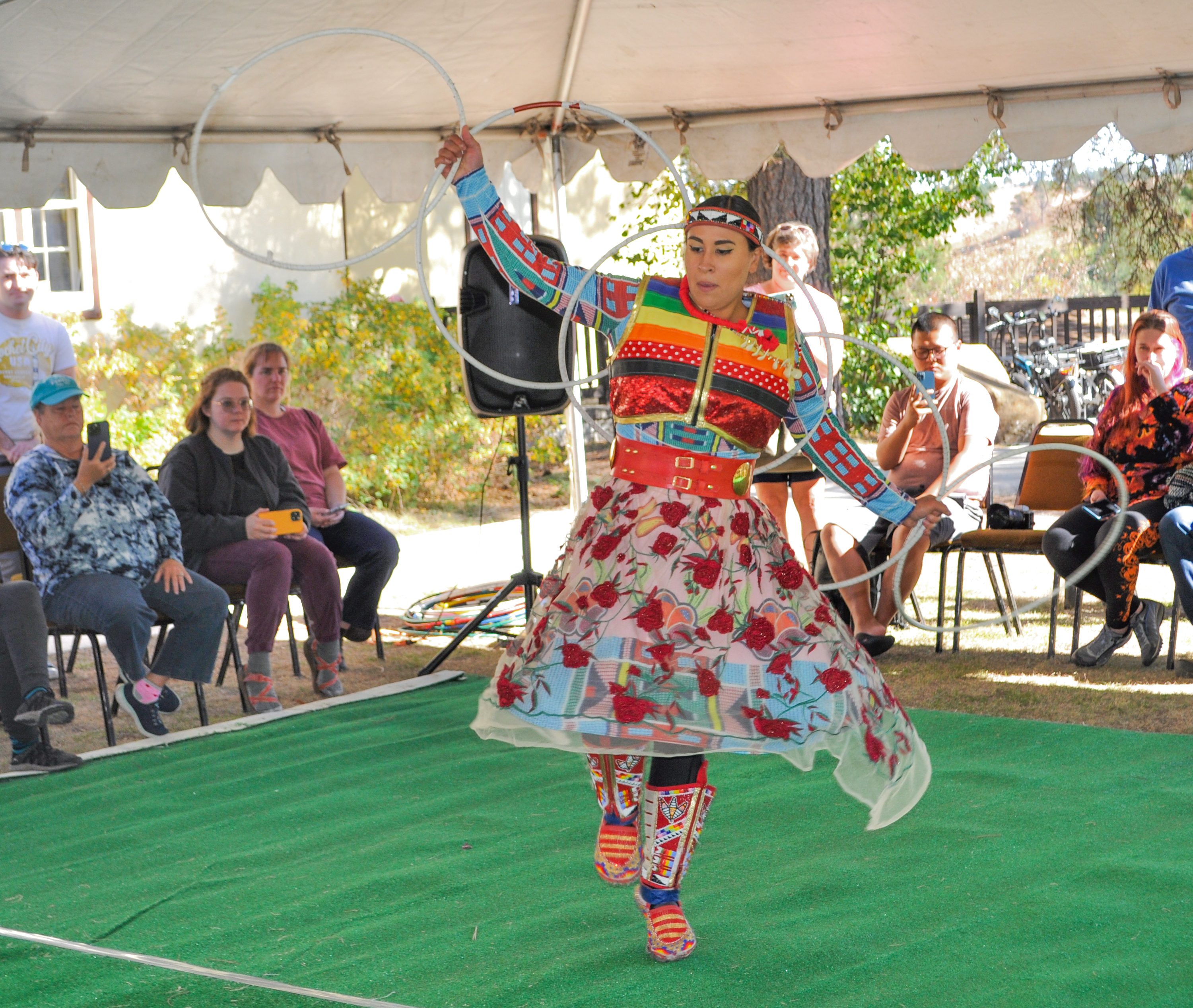Woman wearing a colorful dress dances on a green carpet with white hoops along her arms and in her hands. People stand and sit along the edges of the carpet watching her