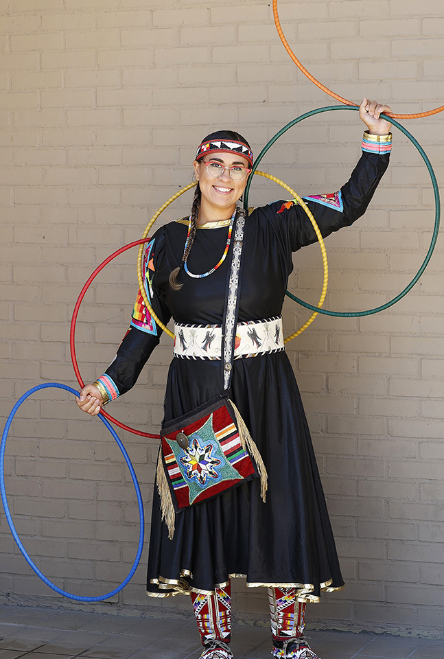 Woman wearing a black dress with beadwork on her dress and moccasins. She is holding four colorful hoops while standing in front of a tan brick wall.