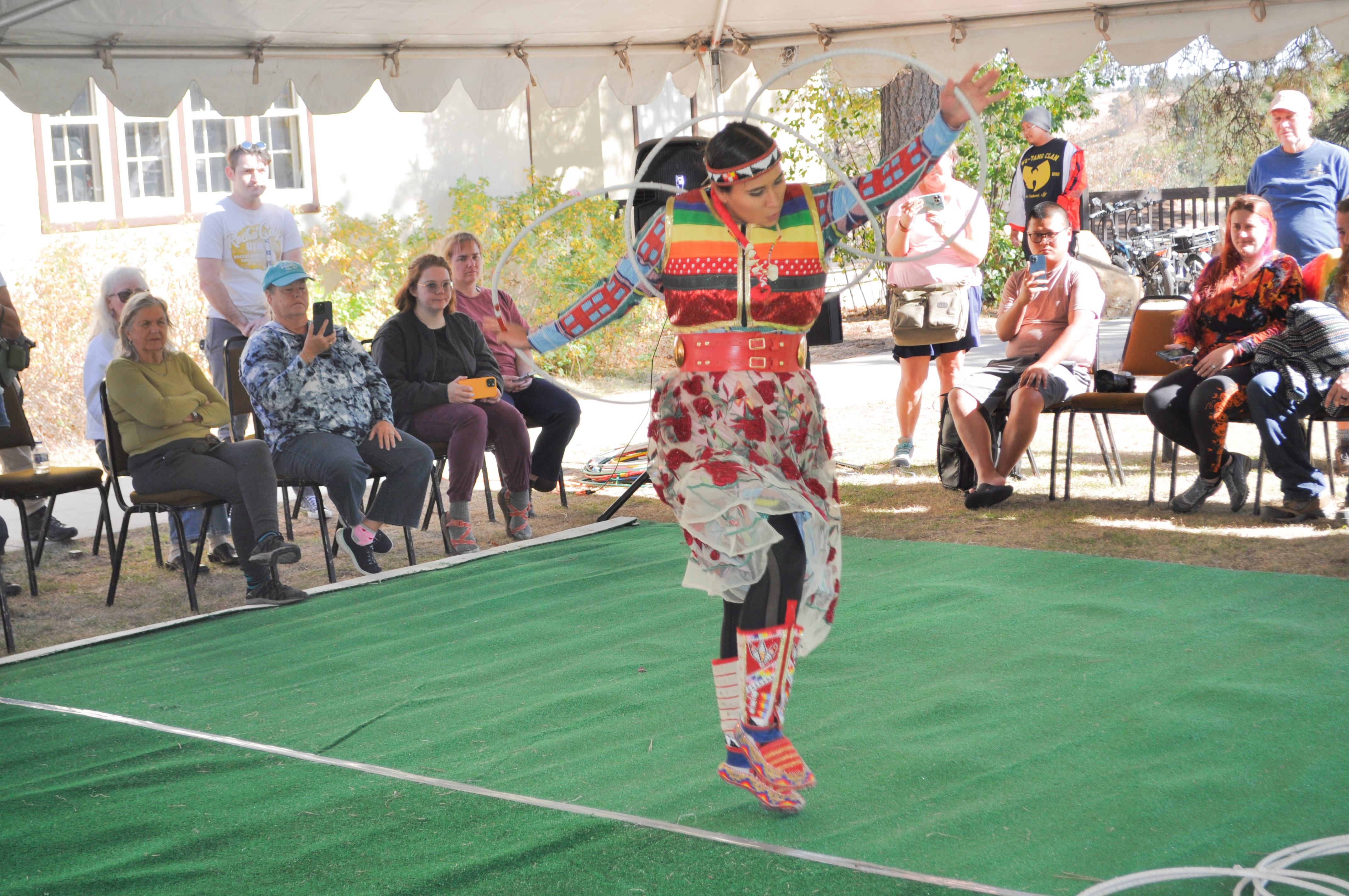 Starr Chief Eagle Woman wearing a colorful dress dances on a green carpet with white hoops along her arms and in her hands. People stand and sit along the edges of the carpet watching her.