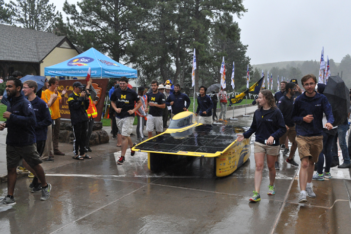 Solar car crossing the finish line surrounded by a crowd of spectators on a wet day.