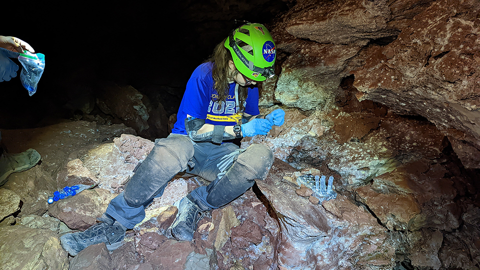 In the center of the photo is a woman sitting in the cave wearing a green caving helmet. She is looking down and wearing blue gloves while holding a small bluish plastic test tube. She is wearing knee pads covered with brown dirt from the cave.