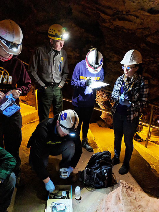 Group of students in the cave, wearing hardhats, gathered in a group as they collect water samples in bottles. Group of students in the cave, wearing hardhats, gathered in a group as they collect water samples in bottles.
