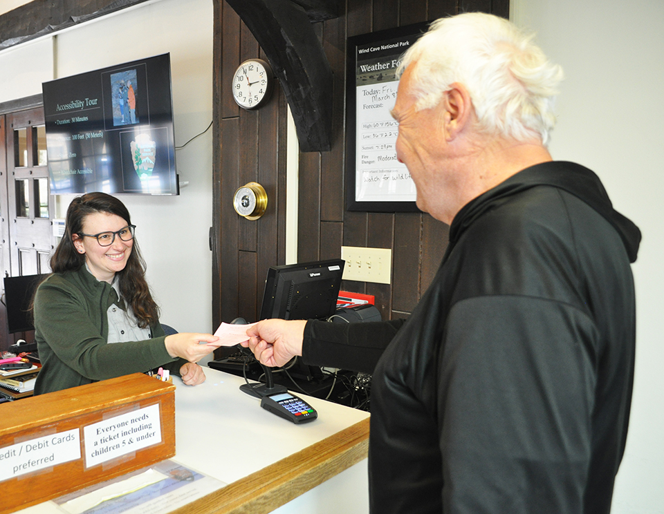 In the center of the picture is a female ranger, standing behind a white information desk with a cash register on it. She is handing a ticket to a man standing on the other side of the information deskr with his back to the camera and wearing a black wind