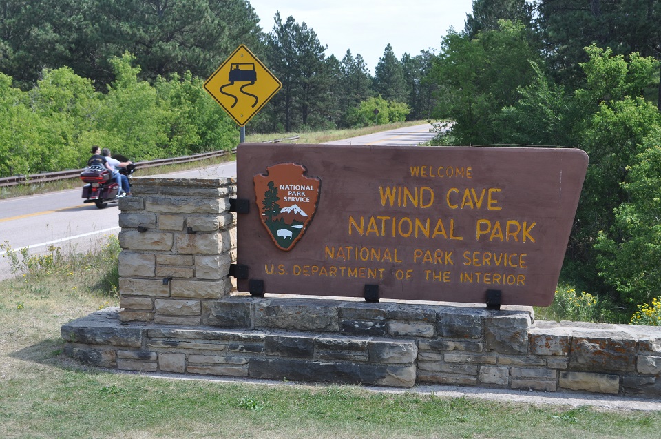 In the middle of the photograph is a large wooden sign on a stone base that welcomes people to Wind Cave National Park. To the left of the sign is a highway with two people riding a motorcycle in the direction of the park. In the middle of the photograph is a large wooden sign on a stone base that welcomes people to Wind Cave National Park. To the left of the sign is a highway with two people riding a motorcycle in the direction of the park.
