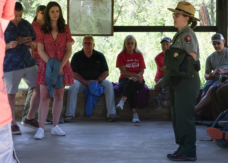 To the right in the photo is a female park ranger talking to a group of people standing or sitting on a rock wall under an outdoor shelter. To the right in the photo is a female park ranger talking to a group of people standing or sitting on a rock wall under an outdoor shelter.