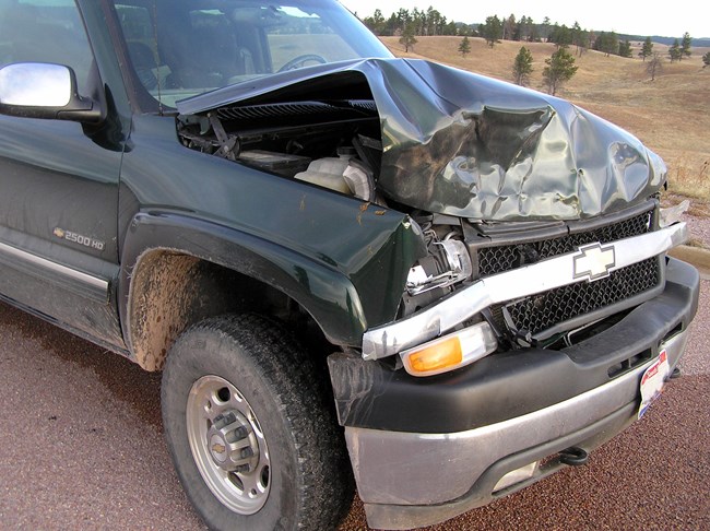 Front end of a green pickup truck with a smashed in grill, broken headlights, and a crumpled hood.